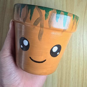 May include: A small, light brown terracotta plant pot featuring a hand-painted face with large black eyes, white highlights, and a simple curved smile. The upper rim is decorated with green and brown painted vine-like patterns.
