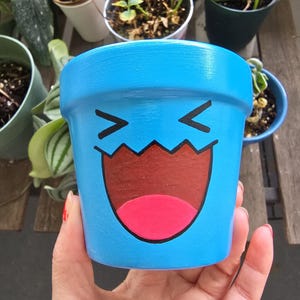 May include: A light blue ceramic plant pot with a cartoon face design. The face features black angled eyebrows, a brown mouth, and a pink tongue. The pot is held in a person's hand, with other potted plants in the background.