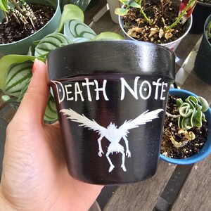 May include: Black painted ceramic flower pot with the words "DEATH NOTE" in white lettering. A white illustration of a winged creature is below the text. The pot is held in a hand.