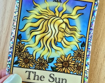Puede incluir: Una pegatina de carta del tarot que representa el Sol. La carta tiene un sol amarillo con una cara, rodeado de girasoles, sobre un fondo azul. La carta tiene un borde plateado brillante y el texto "The Sun".