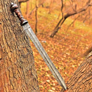 May include: A Damascus steel sword with a wooden handle and a decorative pommel. The blade has a distinctive pattern, and the handle is a combination of dark and light wood tones. The sword is leaning against a tree trunk.