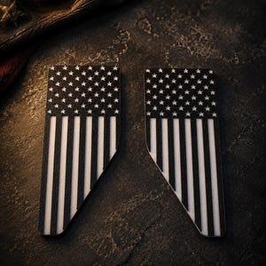 May include: Two rectangular emblems featuring a black and white American flag design. Each emblem has a flat top edge, a vertical left side, and a diagonal cut along the bottom right corner, displaying the stars and stripes in a high-contrast finish.