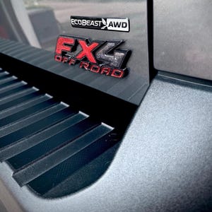 May include: A grey vehicle body panel featuring a black and red FX4 OFF ROAD emblem and a black and white ECOBEAST AWD badge. The surface includes a textured black plastic step component with horizontal ridges along the lower section of the frame.