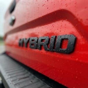 Könnte beinhalten: Eine leuchtend rote Fahrzeugheckklappe mit dem Wort HYBRID in fetten schwarzen Buchstaben. Die glänzende rote Oberfläche ist mit kleinen Wassertropfen bedeckt und ein dunkler, strukturierter Stoßfänger ist am unteren Rand zu sehen.