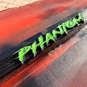 Könnte beinhalten: Eine schwarze Kunststoffschiene mit dem Wort PHANTOM in erhabenen, hellgrünen Buchstaben. Die Schiene ist auf einer Oberfläche mit einem rot-schwarzen Farbverlauf montiert. Die Schrift hat eine texturierte, geschichtete 3D-Druck-Optik.