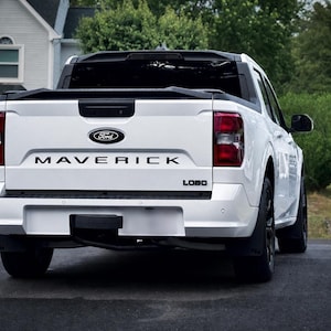 May include: A white Ford Maverick Lobo pickup truck viewed from the rear. The tailgate features a black Ford emblem, the word MAVERICK in bold black lettering, and a small black LOBO badge. The truck has black trim, red taillights, and black wheels.