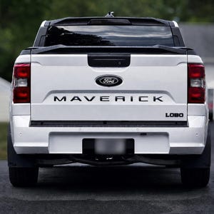 May include: A white Ford Maverick Lobo pickup truck viewed from the rear. The tailgate features the Ford logo in blue and silver, the word MAVERICK in bold black lettering, and the word LOBO in black on the lower right side. Red taillights.