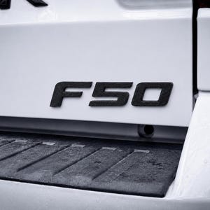 May include: A white vehicle tailgate featuring a matte black F50 emblem centered above a black textured plastic bumper step. The emblem consists of bold, raised lettering with a slightly rough, granular surface texture against the smooth white paint.