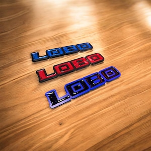 Könnte beinhalten: Drei rechteckige Embleme mit dem Wort LOBO in fetter Blockschrift. Das obere Emblem ist blau, das mittlere rot und das untere schwarz mit blauem Rand. Jedes Teil hat eine glänzende, erhabene Oberfläche und klar definierte Kanten.