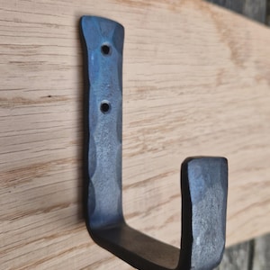 May include: Hand-forged metal hook with a textured, dark blue finish, mounted on a light-coloured wooden board. The hook has a classic J-shape with two drilled holes. Suitable for hanging items in a home or office.