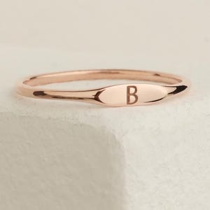 Puede incluir: Anillo de oro rosa con una banda delgada y una cara ovalada. La cara del anillo presenta la letra "B". El anillo se muestra sobre una superficie blanca.