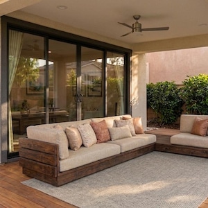 Puede incluir: Conjunto de muebles de patio exterior con un sofá grande de color claro y una sección más pequeña. El sofá tiene un marco de madera oscura y cojines beige, complementados con cojines decorativos. Una alfombra neutra y puertas corredizas de vidrio completan la escena.