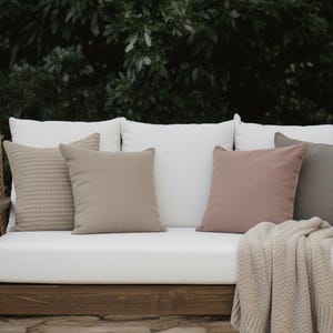 May include: A wooden swing with a white cushion and several throw pillows in shades of beige and pink. A beige blanket is draped over the edge. The swing hangs from thick rope and is set against a backdrop of green foliage.