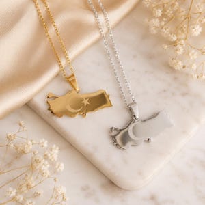 May include: Two necklaces featuring pendants shaped like the map of Turkey, each engraved with a crescent moon and star. One pendant is gold-toned with a matching chain, and the other is silver-toned with a matching chain, both with a smooth finish.
