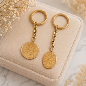 May include: Two gold keyrings with circular and oval pendants. Each pendant has engraved text and hangs from a chain attached to a ring. The keyrings are on a beige surface, with floral accents in the background.