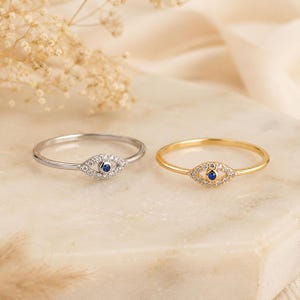 May include: Two thin rings, one silver-coloured and one gold-coloured, each featuring a central eye-shaped motif. The eye design is encrusted with small clear stones surrounding a single, tiny, round blue stone at the centre of each ring band.