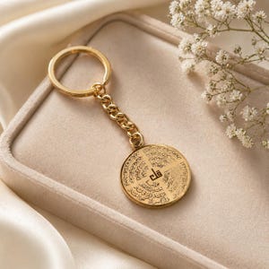 May include: A gold-coloured keyring with a circular pendant. The pendant has detailed engraved text and is attached to a chain and keyring. The background is a soft, neutral colour, with white flowers in the upper right corner.