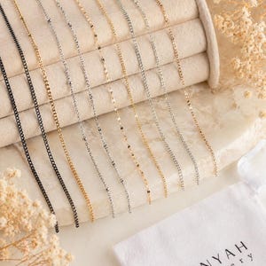 Può includere: Una collezione di delicate collane in varie finiture metalliche, tra cui nero, oro e argento. Le catene presentano diversi stili di maglie, esposte su una superficie beige. È visibile un sacchetto bianco con il marchio "OMNYAH Jewellery".