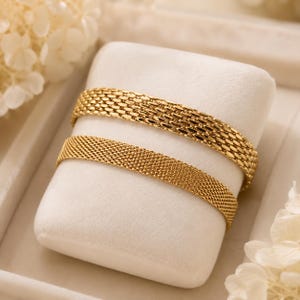 Può includere: Due bracciali color oro esposti su un cuscino di velluto bianco. Il bracciale superiore presenta un motivo a maglie larghe e intrecciate a forma di scaglie, mentre quello inferiore ha una trama a rete più fine e densa con finitura lucida.