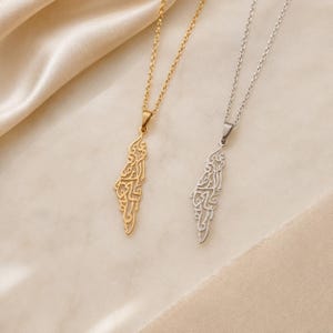 May include: Two necklaces with thin metal chains and pendants shaped like the map of Israel. One pendant is gold-toned and the other is silver-toned. Both pendants feature intricate cut-out Arabic calligraphy designs within the map outline.