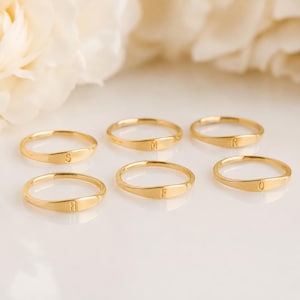 May include: Six gold-coloured rings, each engraved with a different letter: S, M, R, B, F, and Q. The rings are presented on a white surface, with soft, blurred floral elements in the background. The rings have a delicate, minimalist design.