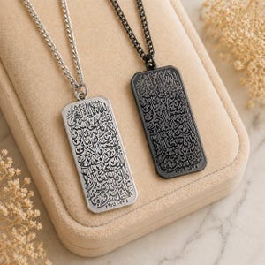 Op de afbeelding: Twee rechthoekige hangerkettingen met ingewikkelde kalligrafie. Eén is zilverkleurig met een zilveren ketting, de andere is zwart met een zwarte ketting. De hangers worden gepresenteerd op een beige oppervlak, dat het gedetailleerde ontwerp laat zien.