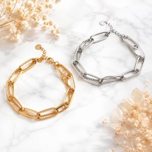 May include: Two layered chain bracelets, one in a gold tone and one in a silver tone. Each bracelet features a combination of large, elongated oval links and a thinner, delicate cable chain. Both bracelets include a small circular charm.