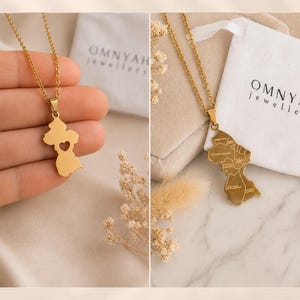 May include: A gold-toned necklace featuring a pendant shaped like the map of Guyana. One side displays a cutout heart shape, while the reverse side shows the engraved regional divisions of the country. The pendant hangs from a delicate gold chain.