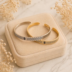 May include: Two open cuff bracelets. One is silver with a black geometric pattern, and the other is gold with a black geometric pattern. The bracelets are displayed on a beige velvet surface, with dried flowers in the background.