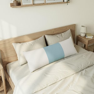 Puede incluir: Una almohada corporal rectangular con un diseño de bloques de color que presenta un panel central ancho de color azul claro flanqueado por dos paneles laterales blancos. La almohada está colocada horizontalmente sobre una cama con ropa de cama clara.