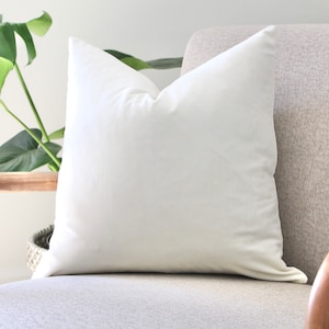 May include: A white pillow on a light gray armchair with wooden arms. The pillow is made of a soft, smooth fabric.