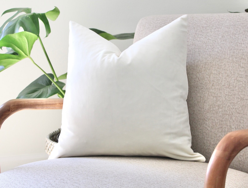 May include: A white pillow on a light gray armchair with wooden arms. The pillow is made of a soft, smooth fabric.