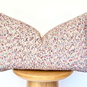 May include: A rectangular decorative pillow with a speckled pattern in various colors, including pink, blue, yellow, and green, on a light background. The pillow is placed on a wooden stool.