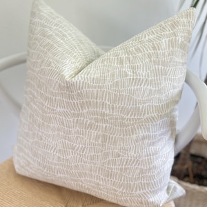 May include: A square decorative pillow with a neutral color palette. The pillow features a light beige fabric with a white, textured, linear pattern. The pillow is placed on a white chair with a woven seat.