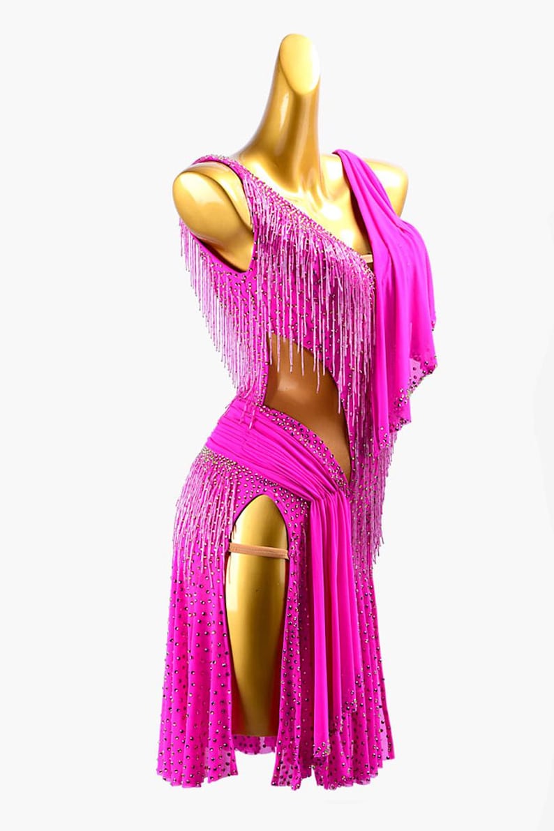 Puede incluir: Un vibrante vestido de baile fucsia con dise&ntilde;o de un solo hombro y abertura alta. El vestido presenta intrincados abalorios y largos flecos. El vestido se exhibe en un maniqu&iacute;.