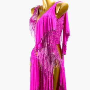 Puede incluir: Un vibrante vestido de baile fucsia con dise&ntilde;o de un solo hombro. El vestido presenta intrincados abalorios y largos flecos. La falda tiene una abertura alta y el vestido se exhibe en un maniqu&iacute; dorado.