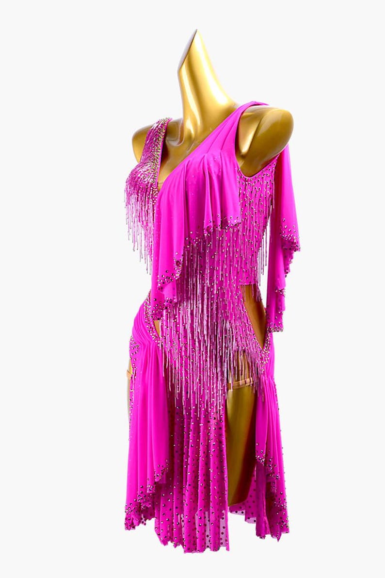 Puede incluir: Un vibrante vestido de baile fucsia con dise&ntilde;o de un solo hombro. El vestido presenta intrincados abalorios y largos flecos. La falda tiene una abertura alta y el vestido se exhibe en un maniqu&iacute; dorado.