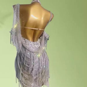 May include: A silver, sleeveless dance dress with a low back and fringe detailing. The dress is embellished with sparkling sequins and hangs on a mannequin. The dress is designed for dance performances.