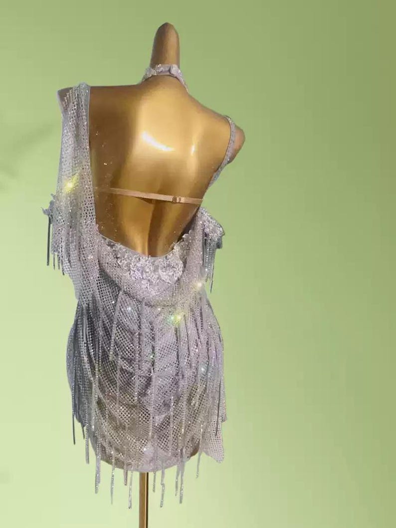 May include: A silver, sleeveless dance dress with a low back and fringe detailing. The dress is embellished with sparkling sequins and hangs on a mannequin. The dress is designed for dance performances.