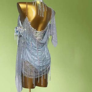 May include: A silver dance dress with intricate rhinestone and fringe detailing. The dress is displayed on a mannequin, showcasing its open back and asymmetrical design. The dress features a halter neckline and a draped, flowing skirt.