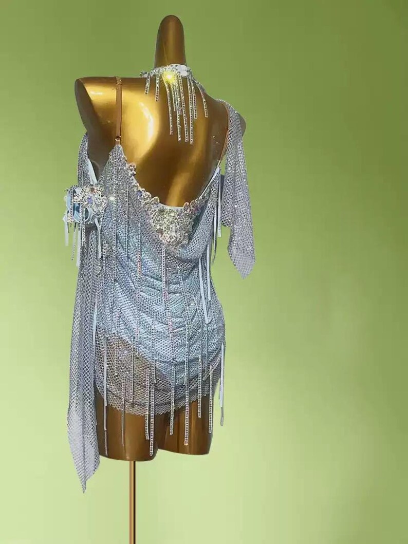 May include: A silver dance dress with intricate rhinestone and fringe detailing. The dress is displayed on a mannequin, showcasing its open back and asymmetrical design. The dress features a halter neckline and a draped, flowing skirt.