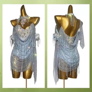 May include: A silver dance dress with intricate rhinestone and fringe detailing. The dress is displayed on a gold mannequin, showcasing its design from both the front and back. The dress features a halter neckline and a draped skirt.