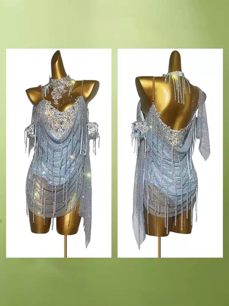 May include: A silver dance dress with intricate rhinestone and fringe detailing. The dress is displayed on a gold mannequin, showcasing its design from both the front and back. The dress features a halter neckline and a draped skirt.