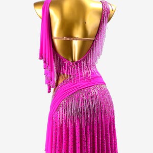 Puede incluir: Un vibrante vestido de baile fucsia con espalda baja y dise&ntilde;o asim&eacute;trico. El vestido presenta flecos de cuentas y adornos brillantes. La falda est&aacute; plisada y adornada con pedrer&iacute;a dispersa. Un detalle de tela drapeada se extiende desde el hombro.