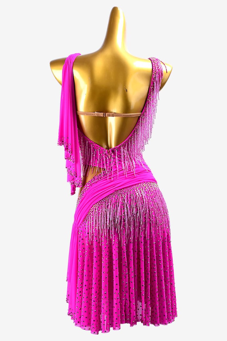 Puede incluir: Un vibrante vestido de baile fucsia con espalda baja y dise&ntilde;o asim&eacute;trico. El vestido presenta flecos de cuentas y adornos brillantes. La falda est&aacute; plisada y adornada con pedrer&iacute;a dispersa. Un detalle de tela drapeada se extiende desde el hombro.