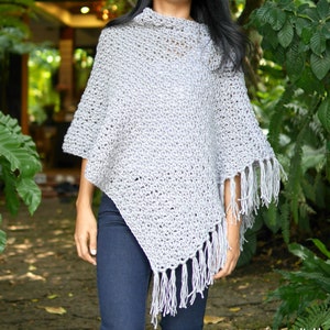 May include: A grey crochet poncho with fringe. The poncho is being worn by a person with long dark hair. The poncho is made using a simple stitch pattern.