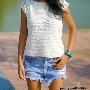 May include: A white crochet top with a loose, open-weave pattern. The top has short, capped sleeves and a round neckline. The model is wearing the top with denim shorts.