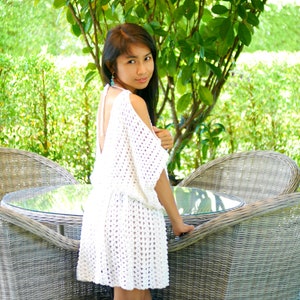 May include: A woman wearing a white crochet dress with a low back and a relaxed fit. She is standing in front of a wicker chair and a table with a glass top.