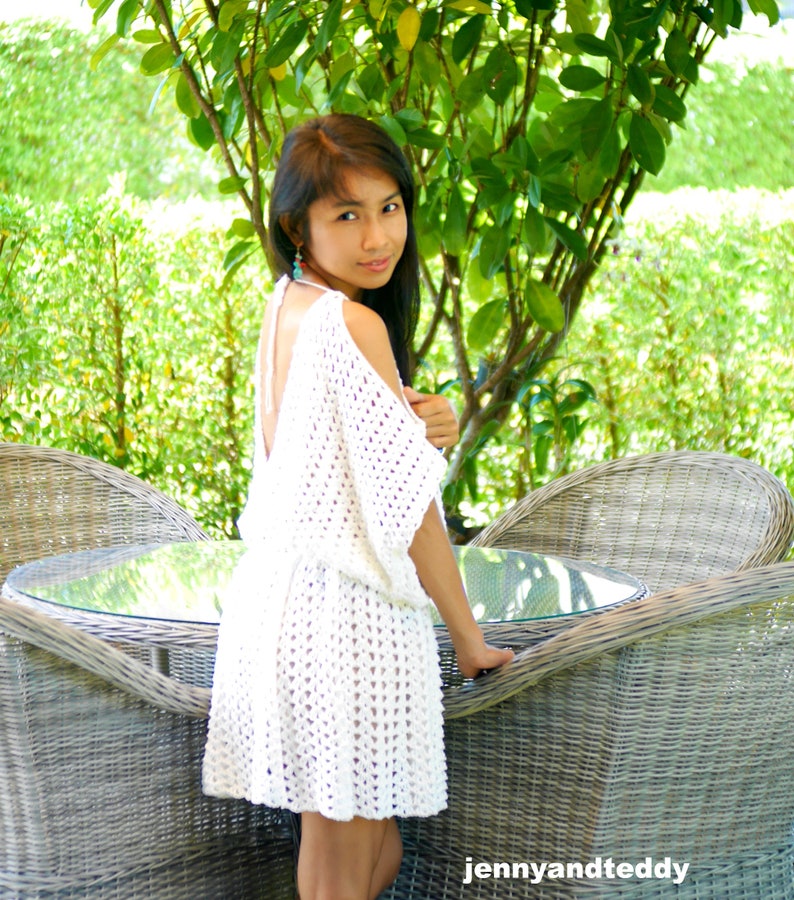 May include: A woman wearing a white crochet dress with a low back and a relaxed fit. She is standing in front of a wicker chair and a table with a glass top.