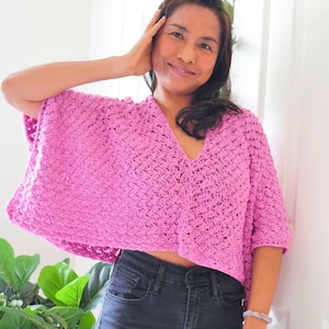 May include: A pink crocheted top with a V-neck and short sleeves. The top is cropped and has a loose fit. The model is wearing the top with black jeans.